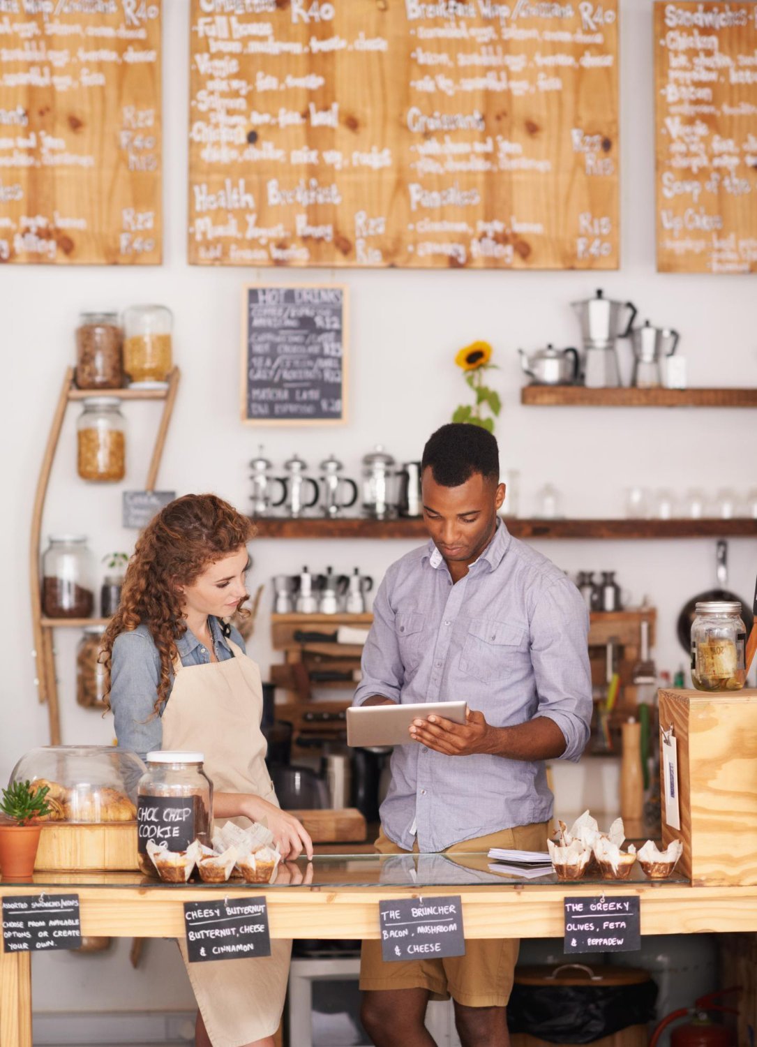 New Hampshire Two people in a coffee shop, one wearing an apron behind the counter and the other holding a tablet, appear to be discussing marketing solutions for small businesses. Menu boards and baked goods are visible in the background. Marketing Agency