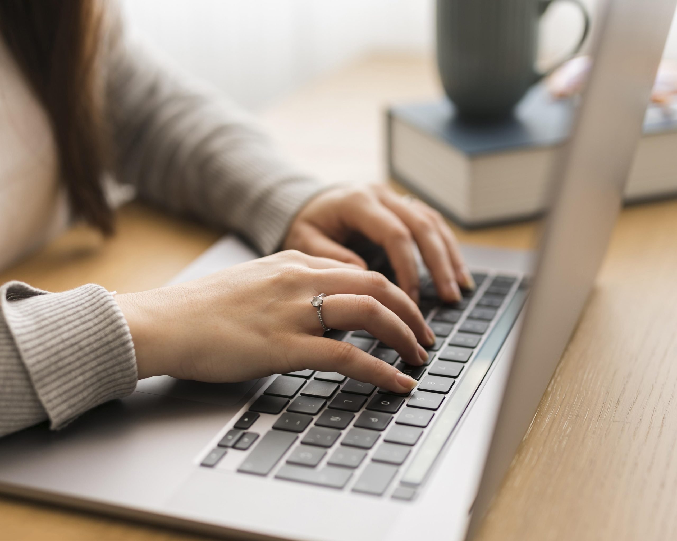Keene Marketing Agency Close-up of a woman's hands typing on a laptop keyboard, with a book on logo design tips and a green mug on the desk.