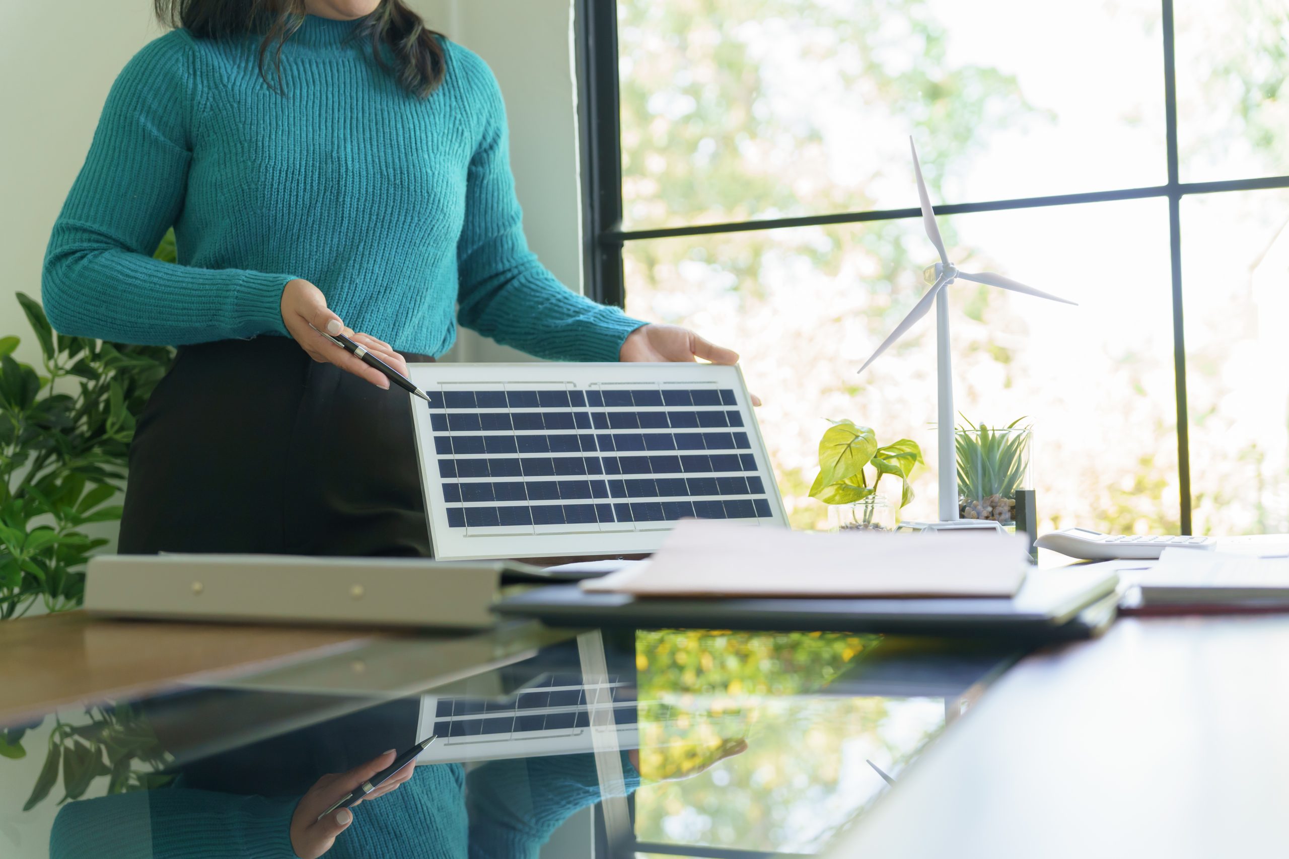 Keene Marketing Agency A woman stands at a desk holding a solar panel, with a small wind turbine model, plants, and laptop nearby in an office with large windows.