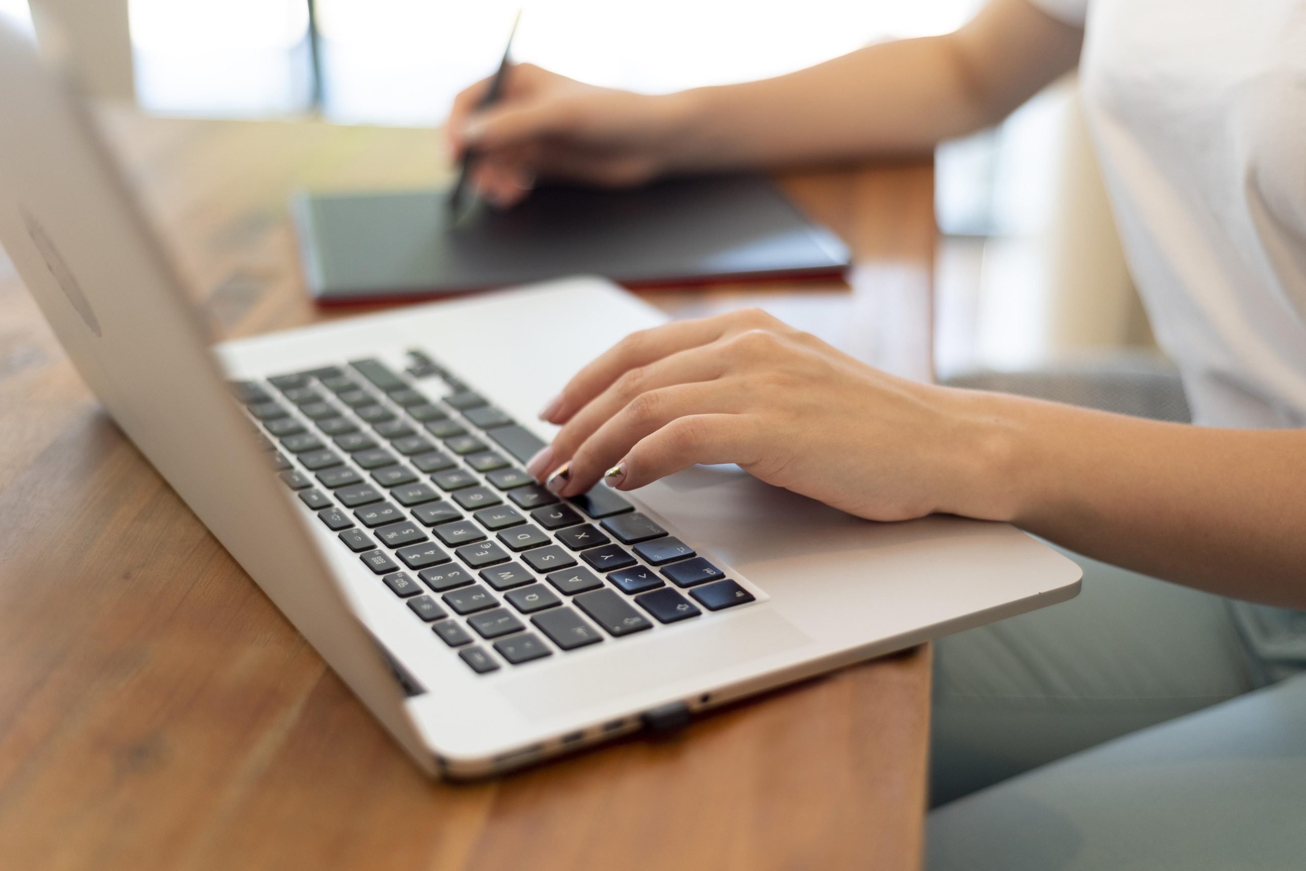 Keene Marketing Agency Close-up of a person's hands typing on a laptop keyboard, with another individual focusing on effective branding strategies in their notebook in the background.