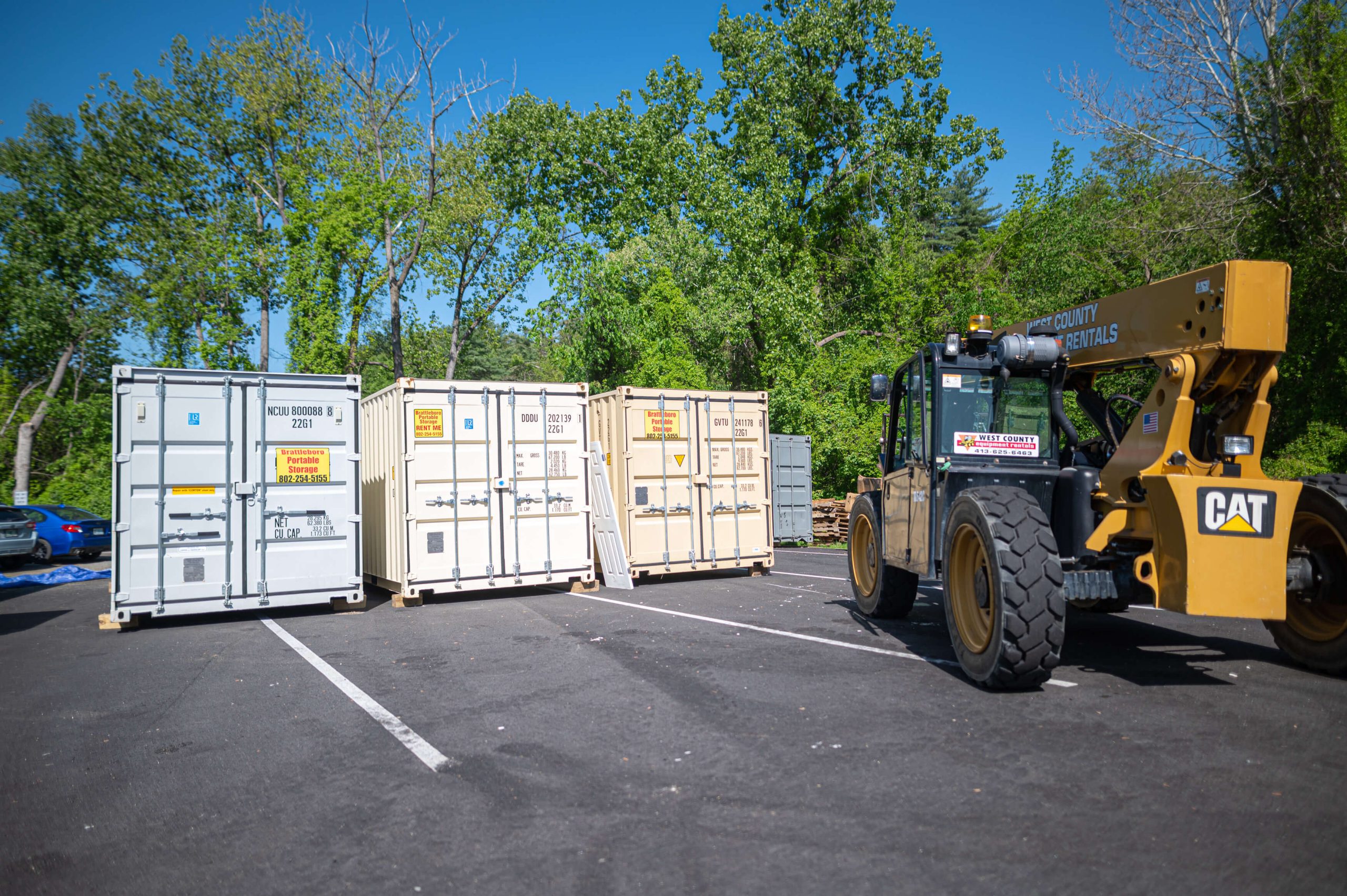 Keene Marketing Agency A forklift labeled "CAT" is parked near four beige shipping containers in a lot surrounded by trees on a clear day.
