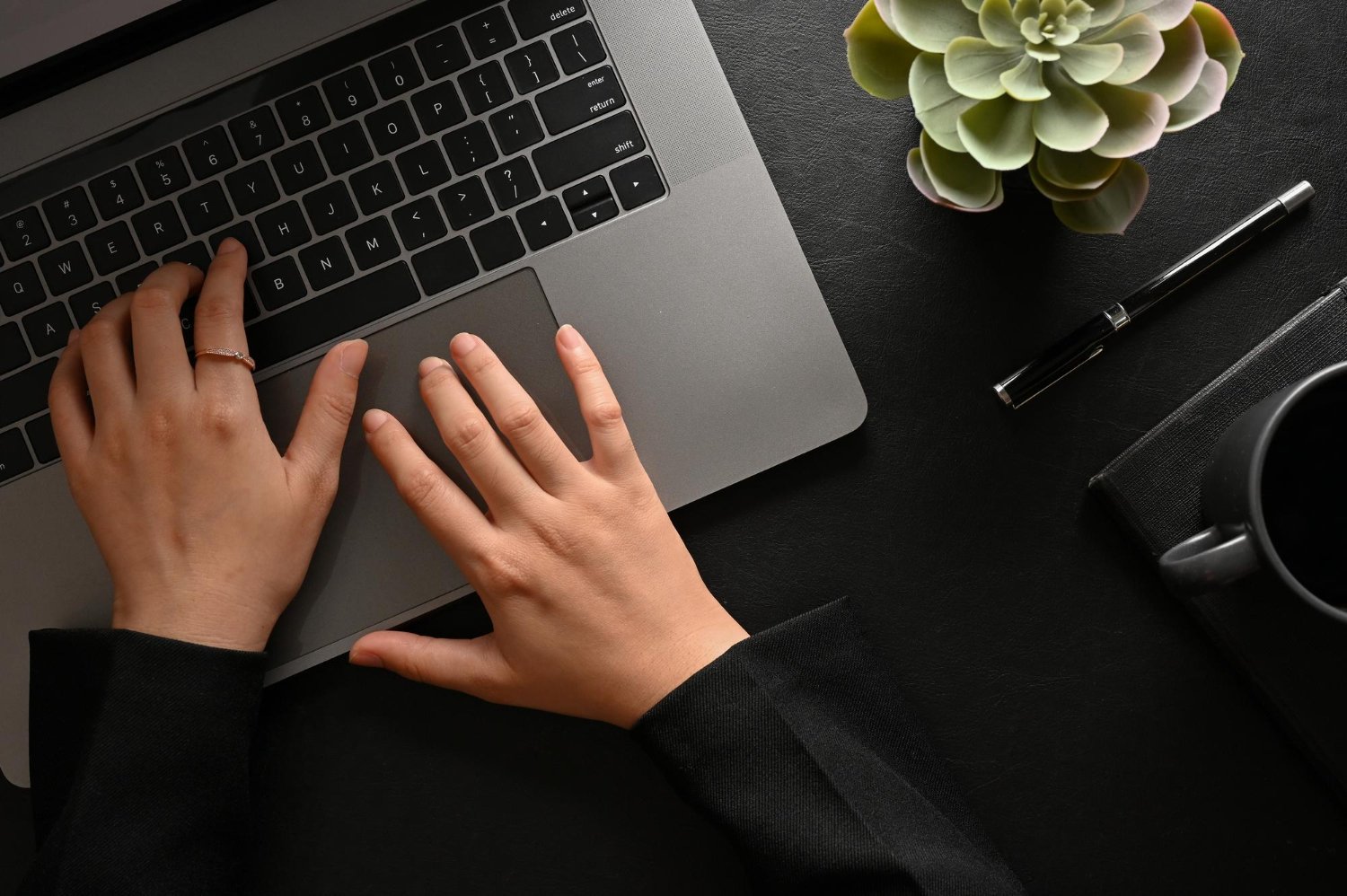 Keene Marketing Agency Hands typing on a laptop keyboard with a succulent plant, pen, black notebook, and coffee mug on a black desk.