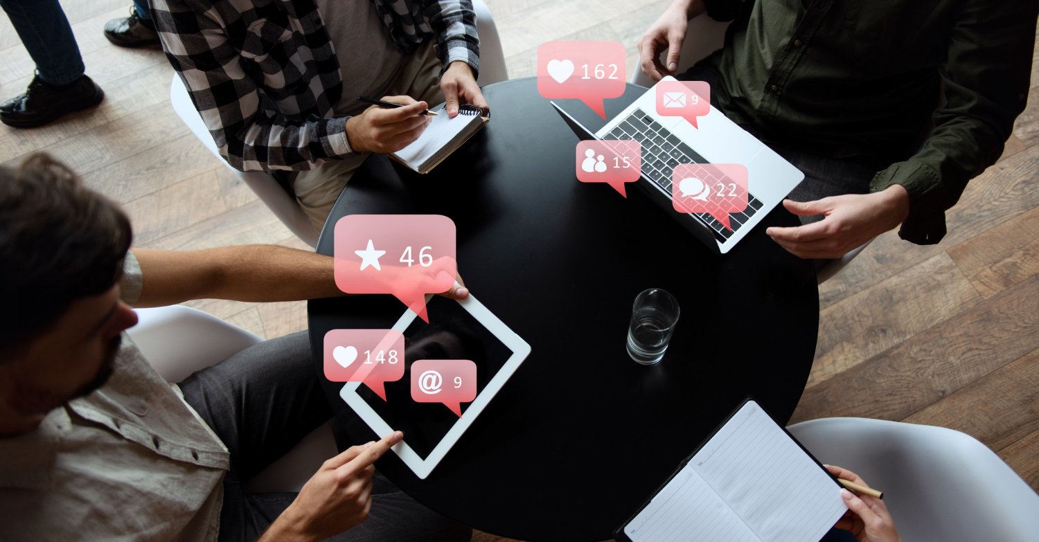 Keene Marketing Agency Four people sit around a round table with electronic devices showing notifications for likes and messages. Notebooks and a glass of water are also on the table.
