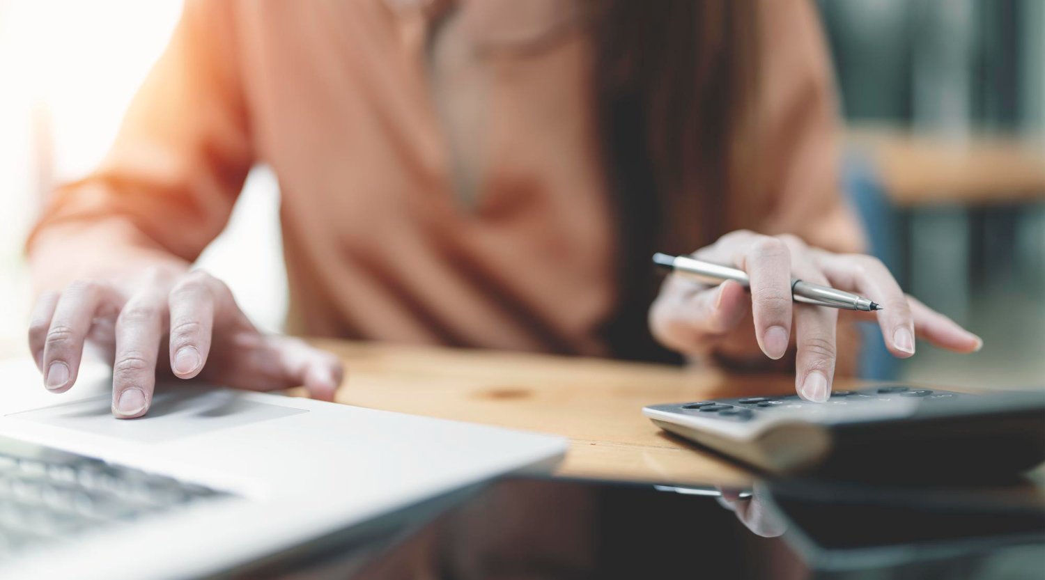 New Hampshire A person in a brown shirt uses a calculator and a laptop on a wooden desk, holding a pen in one hand, concentrating on maximizing their efforts in advertising strategies. Marketing Agency