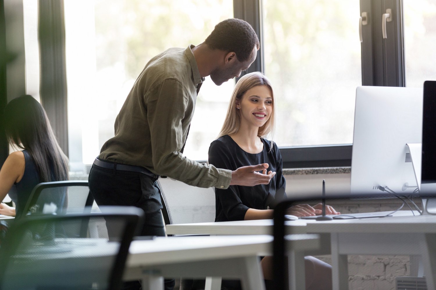 New Hampshire A man and a woman converse at a workstation in a modern office, discussing strategies for success. The man stands while the woman sits at a computer. Additional workers are seen in the background, reflecting a collaborative atmosphere. Marketing Agency