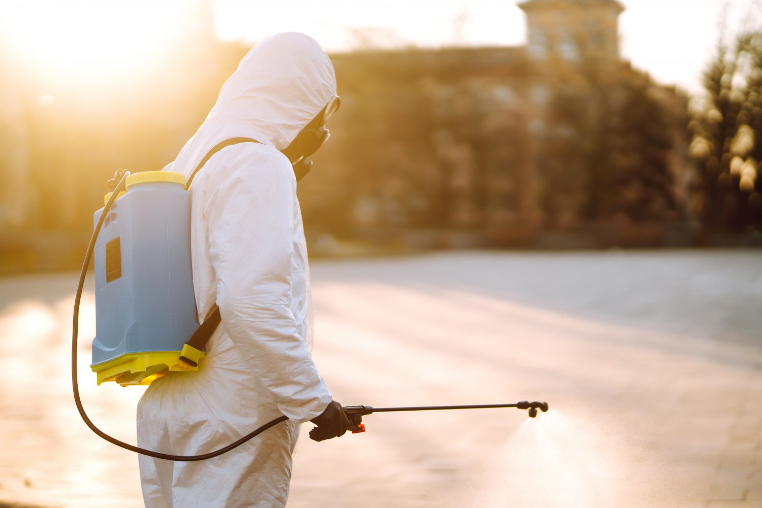 Keene Marketing Agency Person in protective suit and mask spraying disinfectant with a backpack sprayer in an open area at sunset.