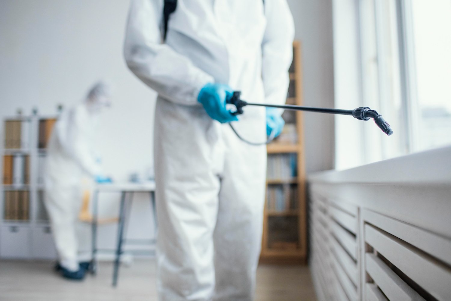 New Hampshire Two people in protective suits and gloves are disinfecting a room; one sprays a substance near a window while the other works in the background, ensuring pest control businesses maintain their standards. Marketing Agency