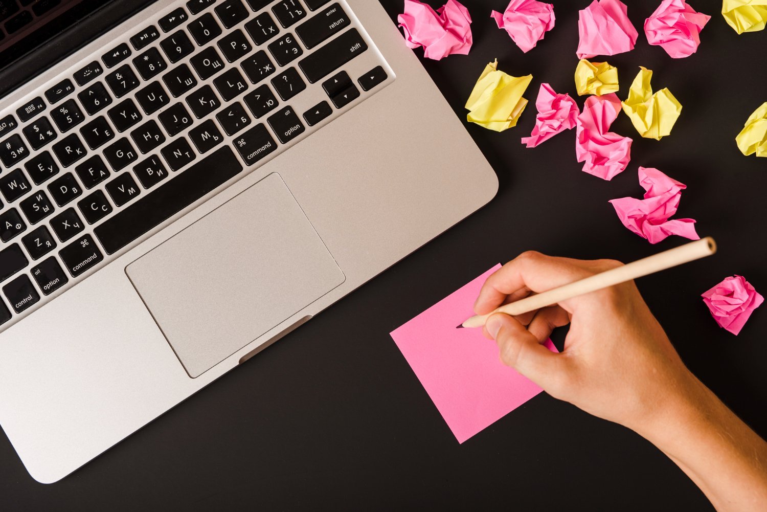 New Hampshire A hand holding a pencil writes on a pink sticky note next to a laptop on a black desk, surrounded by crumpled pink and yellow paper balls—a scene typical in bustling full-service marketing agencies. Marketing Agency
