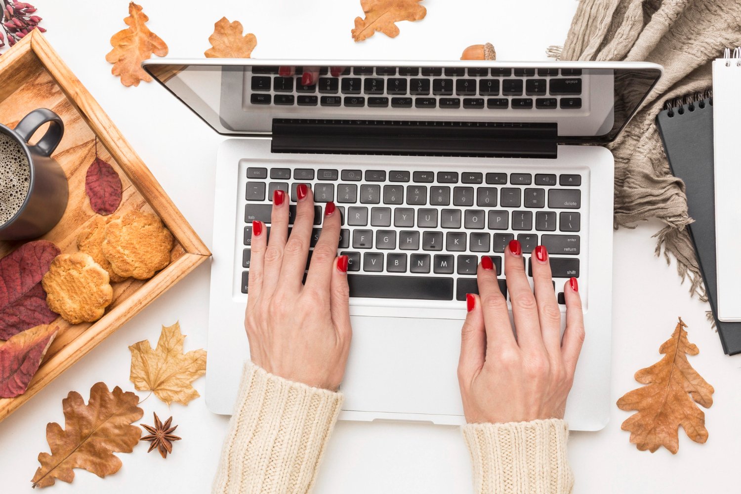 New Hampshire Hands with red nail polish typing on a laptop keyboard surrounded by autumn leaves, a cup of coffee, biscuits, a notepad, and a pen. It's the perfect setting to brainstorm some seasonal marketing tips for home service companies looking to engage their audience during the fall. Marketing Agency
