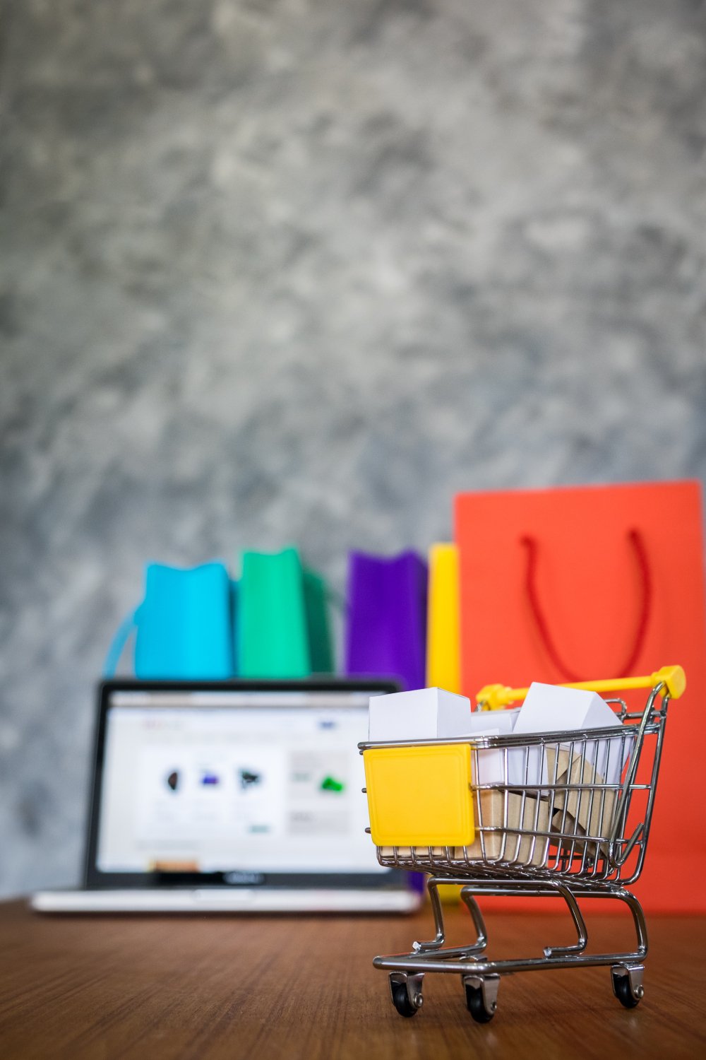 New Hampshire A miniature shopping cart with small boxes sits on a table in front of a laptop displaying a website, embodying a Beginners Guide to retail marketing. Colorful shopping bags are in the background, reflecting strategies for boosting your business. Marketing Agency
