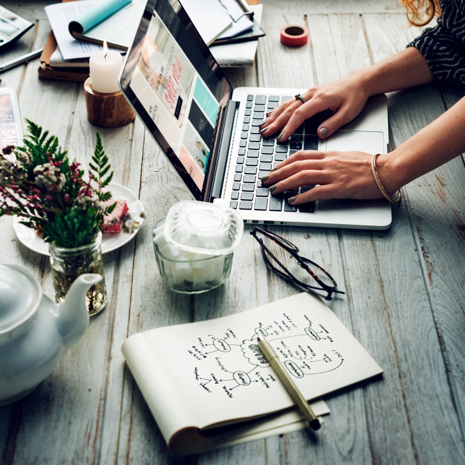 New Hampshire A person types on a laptop at a wooden desk, surrounded by a notebook, pen, teapot, glasses, plants, a lit candle, and various documents—a scene reminiscent of a Vermont business atmosphere focusing on local SEO. Marketing Agency