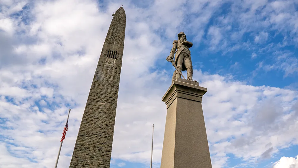 New Hampshire A tall stone obelisk monument stands beside a statue of a man on a pedestal, under a partly cloudy sky with an American flag visible in the background. Marketing Agency