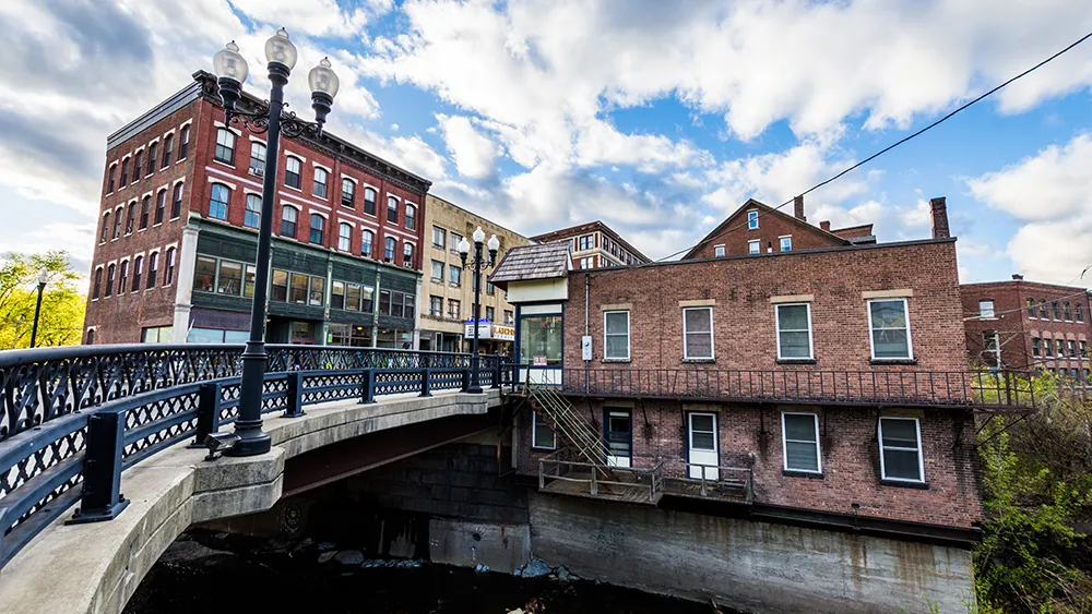 New Hampshire A bridge with a decorative rail leads to a cluster of multi-story red brick buildings under a partly cloudy sky. Marketing Agency