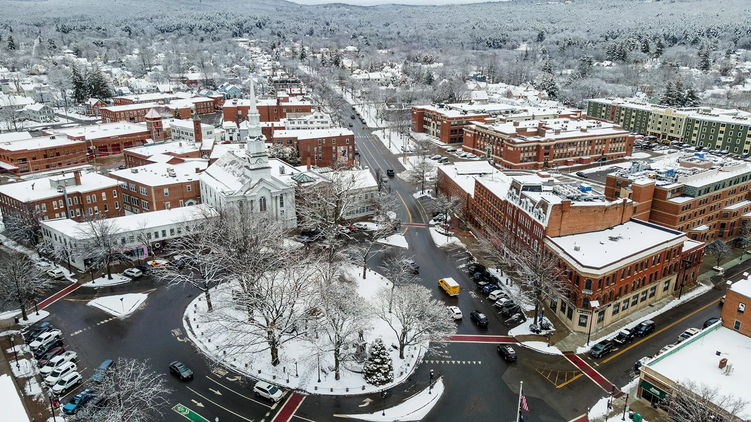 New Hampshire Aerial view of a snow-covered town center with brick buildings, a white church, and a circular road. Snow blankets rooftops and trees, with cars parked along the streets and minimal traffic. Marketing Agency