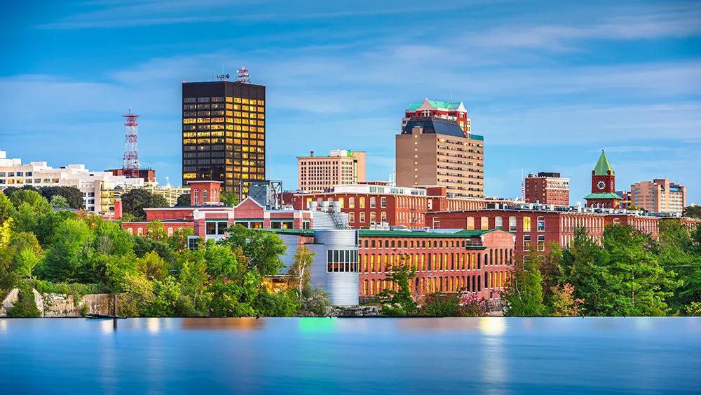 New Hampshire Skyline of Manchester, New Hampshire, featuring a mix of historic mill buildings and modern skyscrapers against a clear blue sky, with a river in the foreground. Marketing Agency