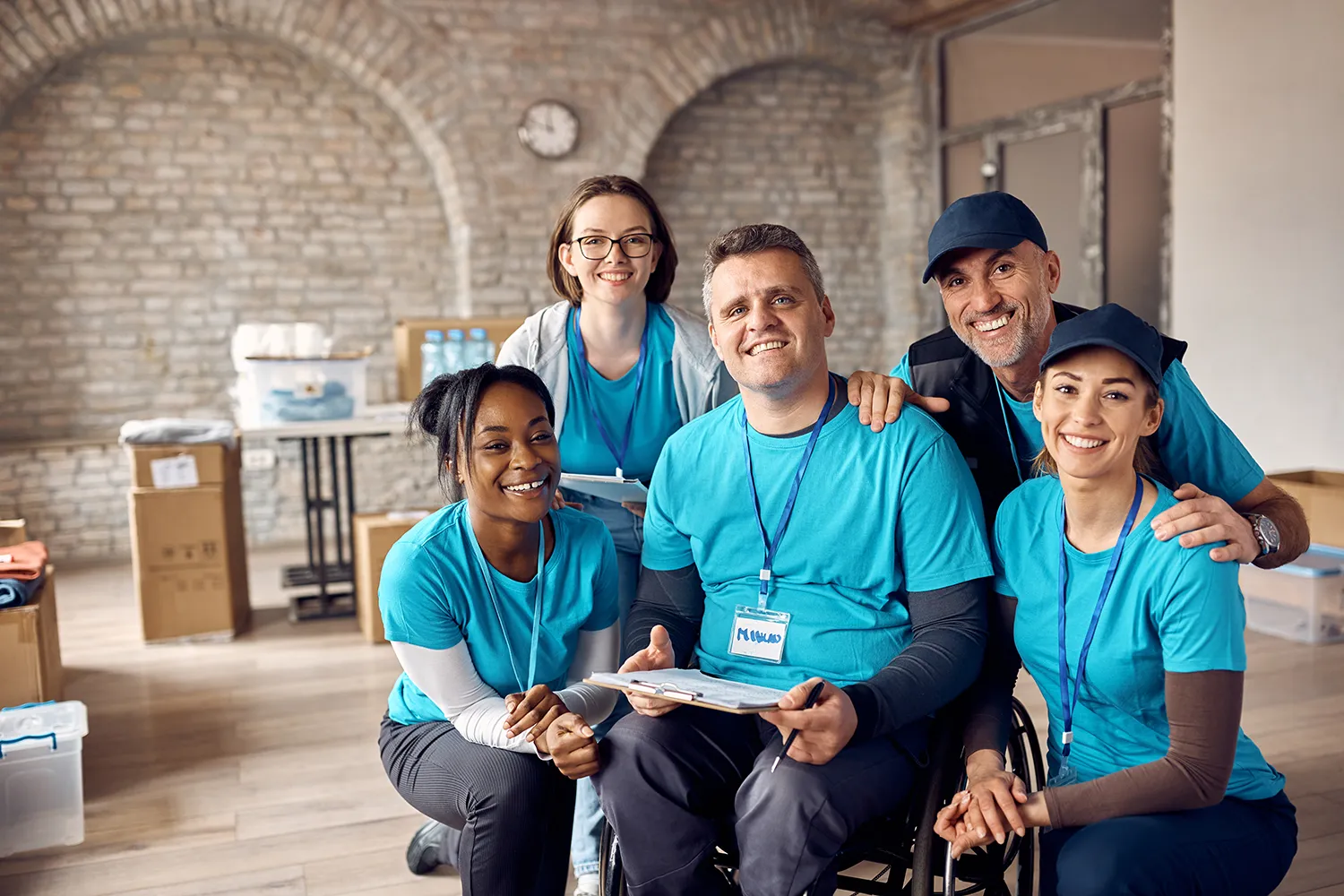 New Hampshire A diverse group of five people, wearing matching blue shirts, smile for the camera in a brick-walled room filled with boxes and supplies, highlighting their teamwork in a nonprofit. One person is seated in a wheelchair. Marketing Agency