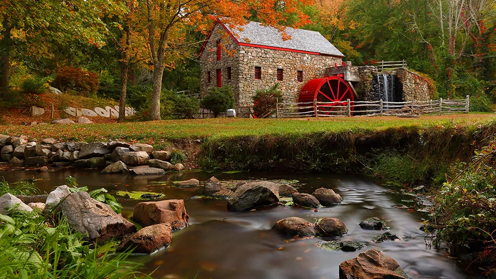 New Hampshire A historic stone mill with a red water wheel stands beside a flowing stream surrounded by lush greenery and autumn trees. Marketing Agency
