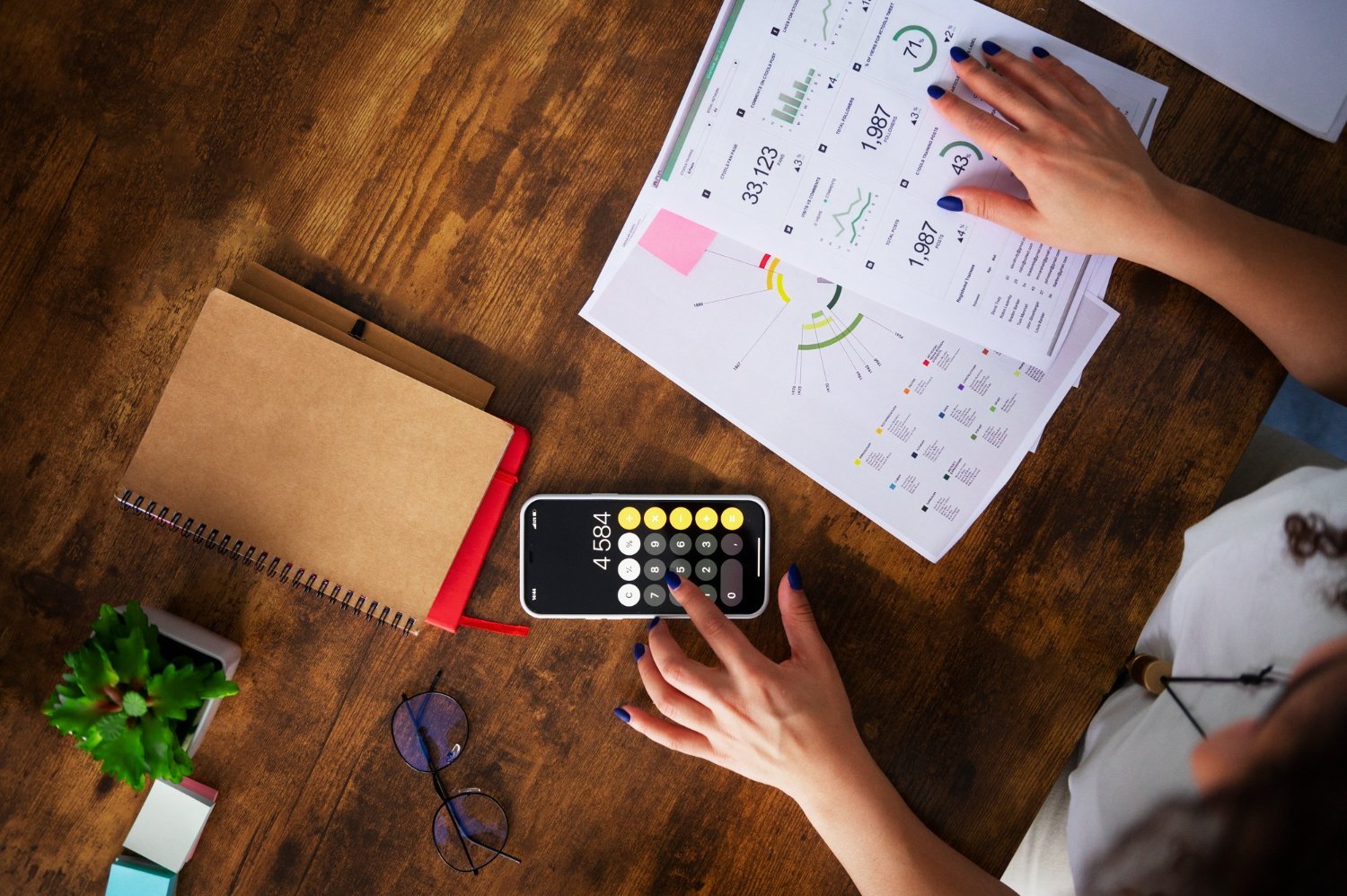 New Hampshire Person using a calculator on their phone while reviewing financial charts and graphs on paper, with a notebook, glasses, and a small plant nearby on a wooden table. They're incorporating budget-friendly tips for small businesses into their analysis. Marketing Agency