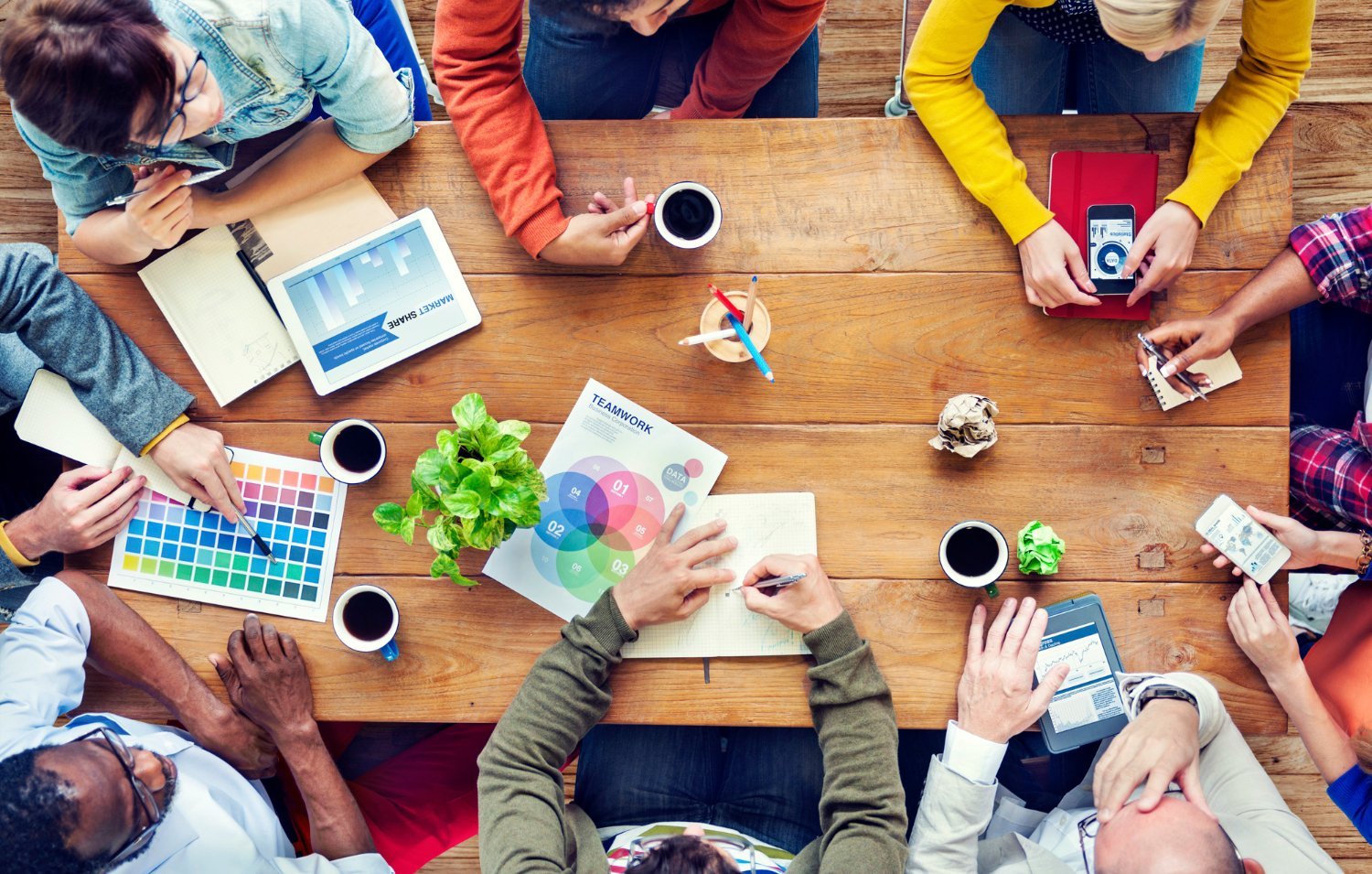 New Hampshire A group of people sitting around a wooden table collaborating with various devices and papers, including a color chart, a plant, a tablet, and coffee cups. They discuss how to hire a marketing agency while considering different marketing agency pricing options. Marketing Agency