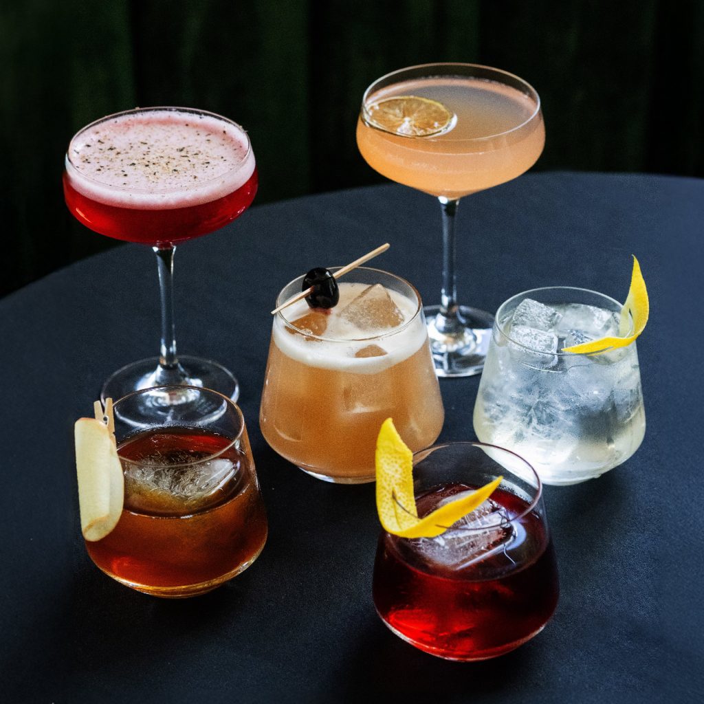 New Hampshire A selection of five cocktails graces a round table at our restaurant, each in a distinct style of glass. They are garnished with citrus, cherries, a cinnamon stick, and ice—perfect for showcasing innovative local marketing strategies that capture the essence of flavor. Marketing Agency