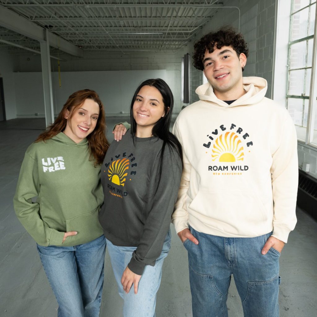 New Hampshire Three people standing indoors wearing hoodies emblazoned with "Live Free" and "Roam Wild," showcasing a proven strategy in apparel that resonates with the adventurous spirit. Marketing Agency