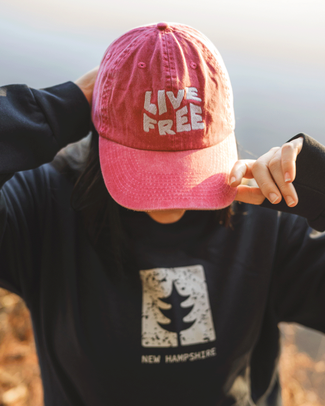 New Hampshire Person wearing a pink hat with "LIVE FREE" text and a dark sweatshirt featuring a tree and "NEW HAMPSHIRE" text. Marketing Agency