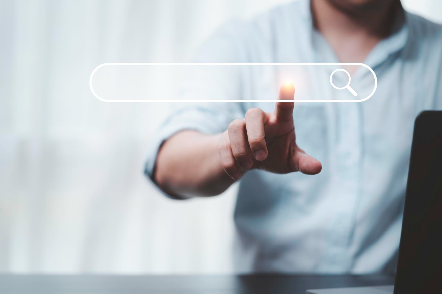New Hampshire A person in a light shirt touches a virtual Google Ads search bar icon on a transparent screen, with a blurred background and part of a laptop visible—highlighting the importance of using a good agency to avoid wasting money. Marketing Agency