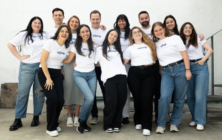 New Hampshire A group of thirteen people standing close together, smiling, and wearing matching white t-shirts and casual pants, posing outdoors against a white wall. Marketing Agency