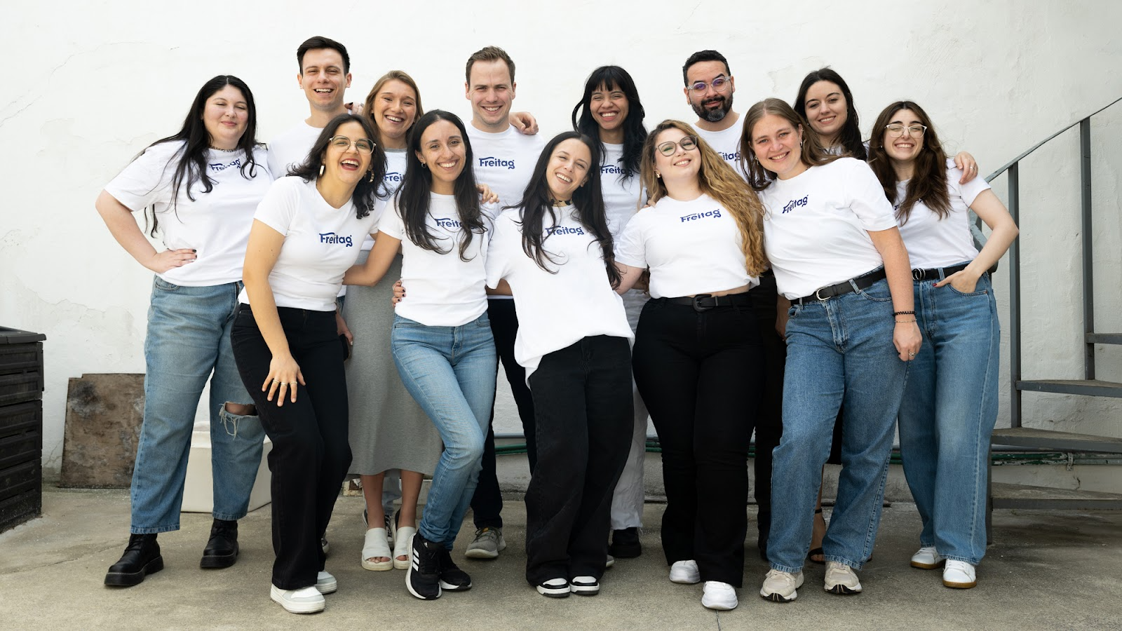 New Hampshire Thirteen people wearing white t-shirts and jeans stand together outdoors, smiling at the camera in front of a white wall and metal stairs. Marketing Agency