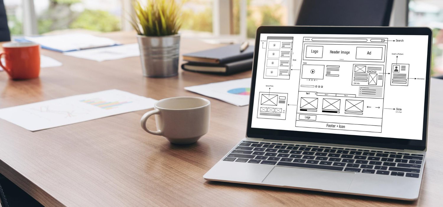 New Hampshire A laptop displaying a good website design wireframe sits on a wooden desk with a coffee cup, papers, and office supplies in a bright workspace—perfect for building good business ideas. Marketing Agency
