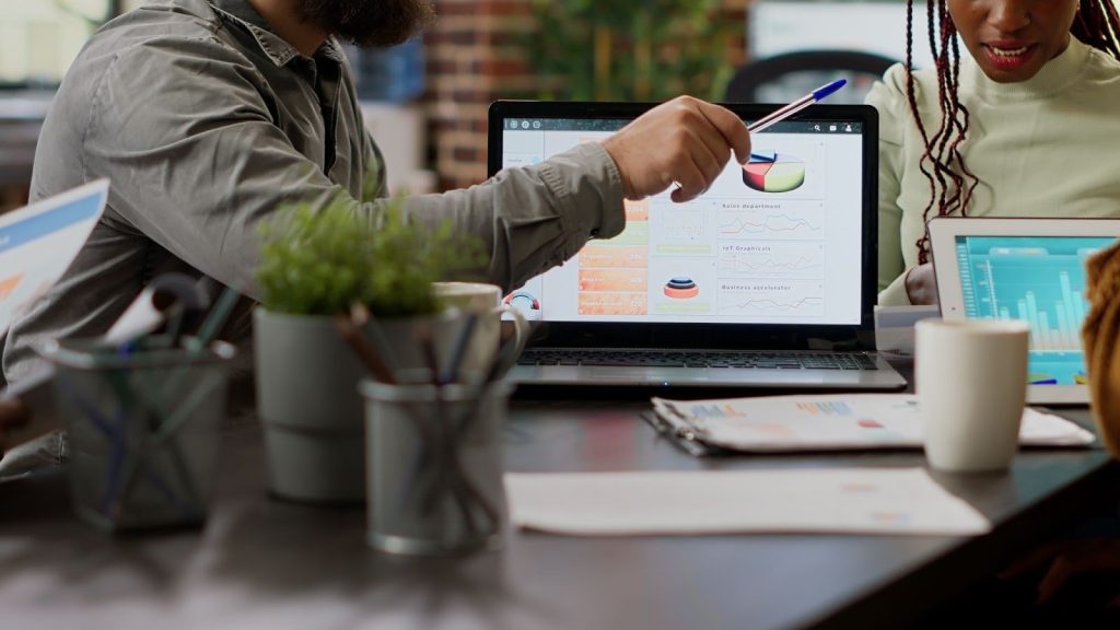 New Hampshire People sitting at a table review charts and graphs displayed on a laptop screen during a meeting; documents and plants are on the desk. Marketing Agency