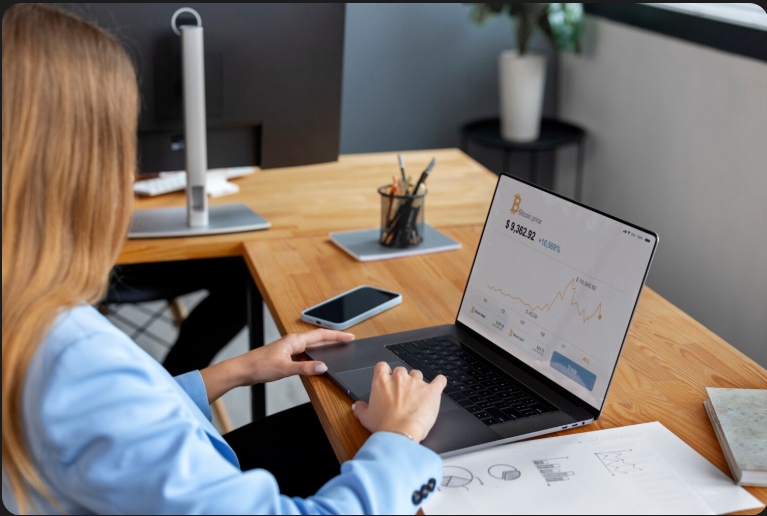 New Hampshire A woman in a blue blazer uses a laptop displaying a Bitcoin price chart at a wooden desk with office supplies and documents. Marketing Agency