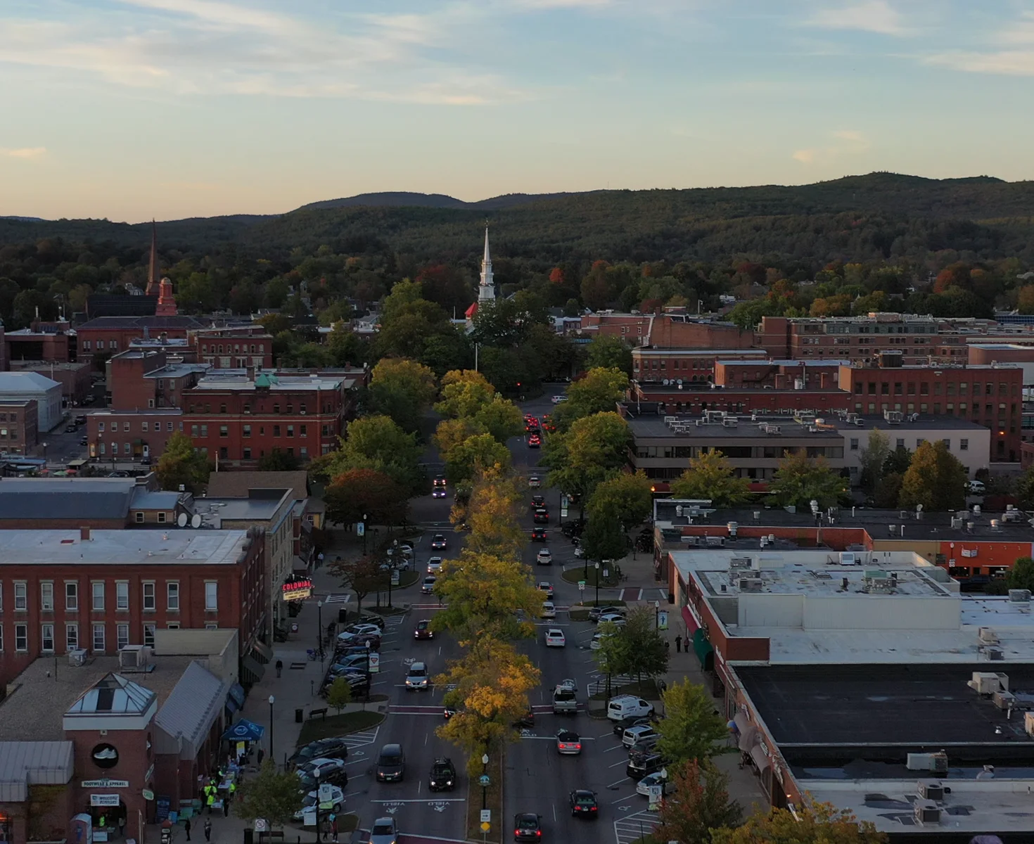 New Hampshire Aerial view of a small town with brick buildings, tree-lined streets, and cars parked along the road, with hills and forest in the background under a partly cloudy sky. Marketing Agency