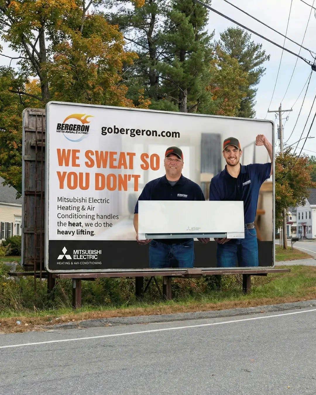 New Hampshire Two men in work uniforms stand in front of a billboard advertising Mitsubishi Electric Heating & Air Conditioning, holding an HVAC unit and smiling. The text reads, "We sweat so you don't. Marketing Agency