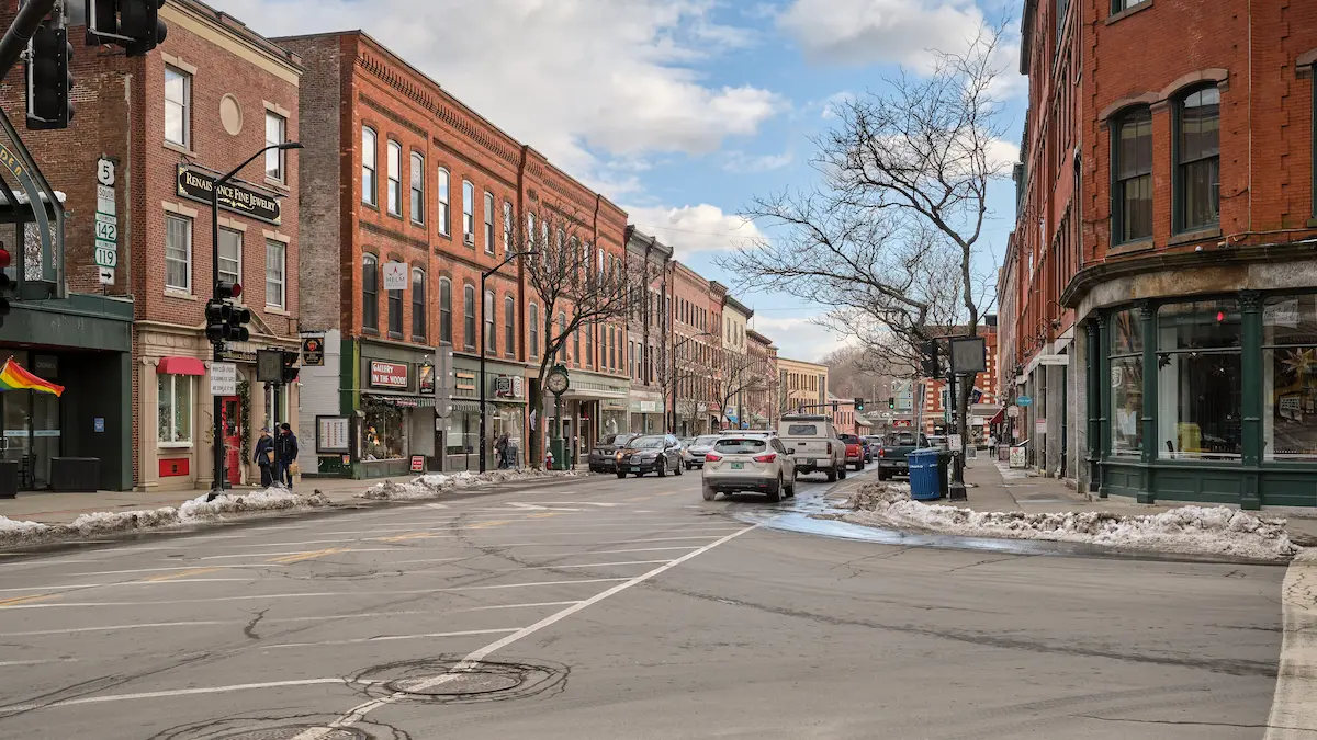 New Hampshire Aerial view of a small town with brick buildings, tree-lined streets, and cars parked along the road, with hills and forest in the background under a partly cloudy sky. Marketing Agency