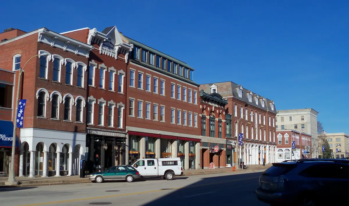 New Hampshire Aerial view of a small town with brick buildings, tree-lined streets, and cars parked along the road, with hills and forest in the background under a partly cloudy sky. Marketing Agency
