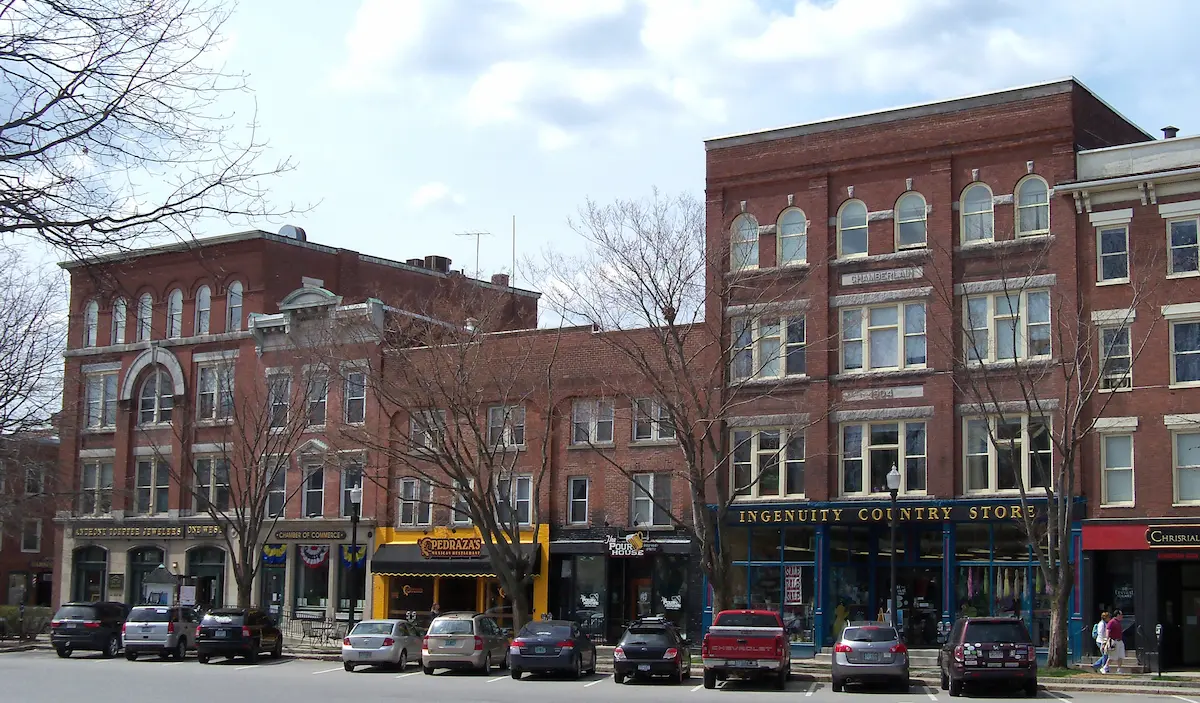 New Hampshire Aerial view of a small town with brick buildings, tree-lined streets, and cars parked along the road, with hills and forest in the background under a partly cloudy sky. Marketing Agency