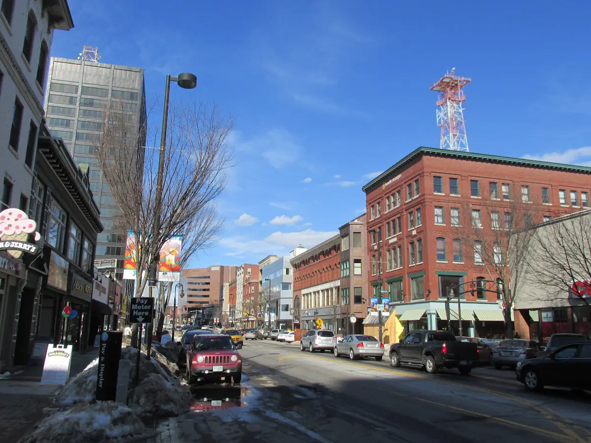 New Hampshire Aerial view of a small town with brick buildings, tree-lined streets, and cars parked along the road, with hills and forest in the background under a partly cloudy sky. Marketing Agency