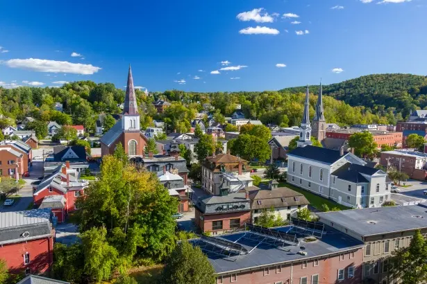 New Hampshire Aerial view of a small town with brick buildings, tree-lined streets, and cars parked along the road, with hills and forest in the background under a partly cloudy sky. Marketing Agency
