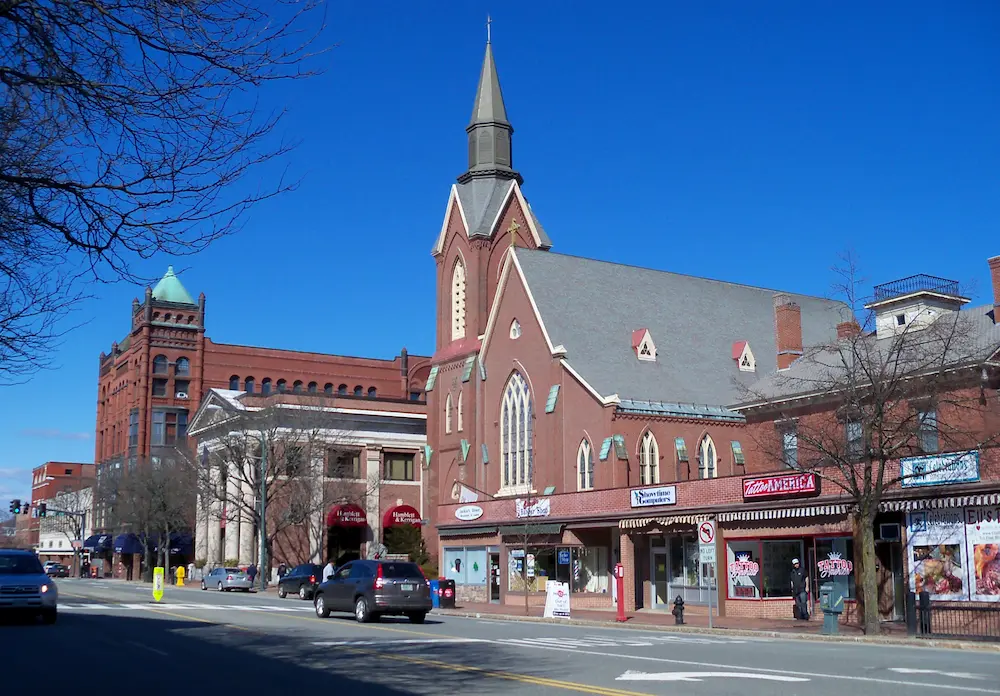 New Hampshire Aerial view of a small town with brick buildings, tree-lined streets, and cars parked along the road, with hills and forest in the background under a partly cloudy sky. Marketing Agency