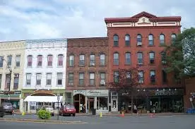 New Hampshire Aerial view of a small town with brick buildings, tree-lined streets, and cars parked along the road, with hills and forest in the background under a partly cloudy sky. Marketing Agency